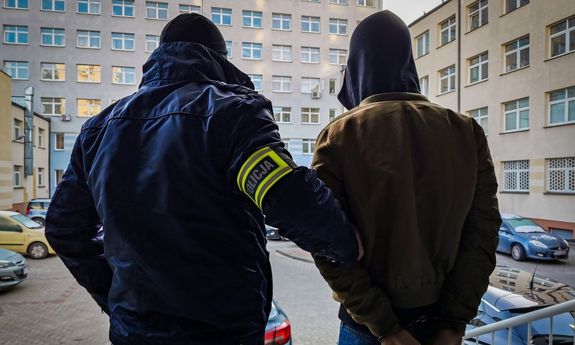 Policjant kryminalny z żółtą, odblaskową opaską na ramieniu z napisem: policja. Policjant prowadzi zatrzymanego mężczyznę, który ma założone kajdanki na ręce trzymane z tyłu