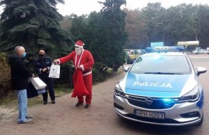 Mikołaj wraz z policjantką przekazują paczki dla dzieci ordynatorowi obok policyjnego radiowozu przed Szpitalem Powiatowym w Kozienicach&quot;&gt;
