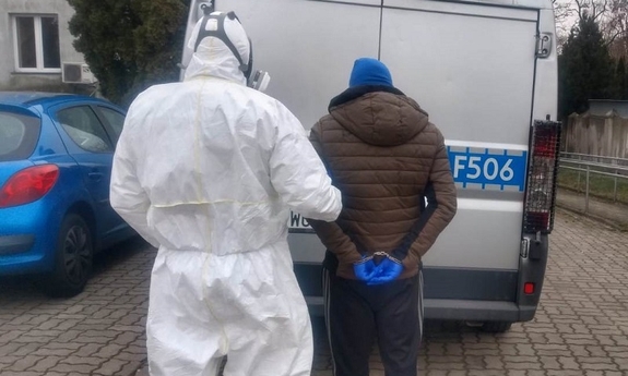 Policjant w kombinezonie ochronnym koloru białego prowadzi zatrzymanego do radiowozu. Przed nimi stoi zaparkowany radiowóz. Zatrzymany ma założone na ręce trzymane z tyłu kajdanki. W tle widoczny budynek. Po lewej stronie widać niebieski samochód osobowy