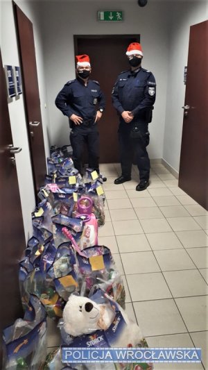 Zdjęcie przedstawia policjantów stojących na korytarzu komendy. Funkcjonariusze są w granatowych mundurach, a na głowach mają czerwone czapki. Obok nich stoją paczki z prezentami