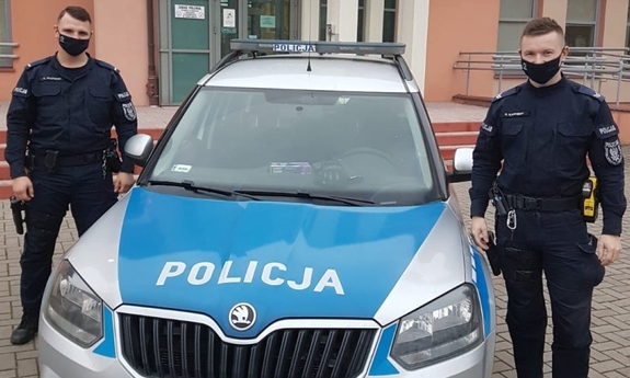 dwaj policjanci stoją obok radiowozu, w tle budynek komendy Policji
