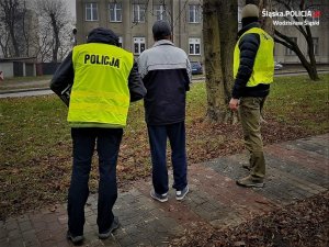 dwaj policjanci w kamizelkach z napisem Policja, prowadzą zatrzymanego mężczyznę - widok z tyłu