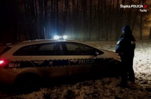 Policjant stoi obok radiowozu, patrzy w stronę lasu, jest ciemno