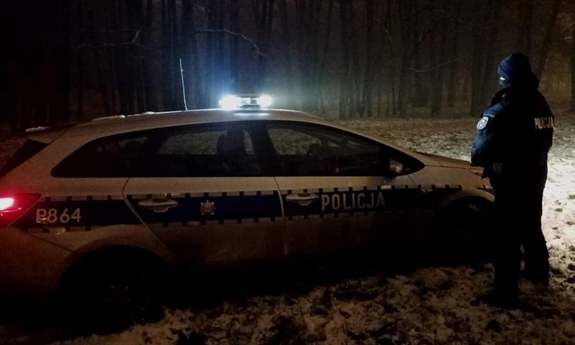 Policjant stoi obok radiowozu, patrzy w stronę lasu, jest ciemno