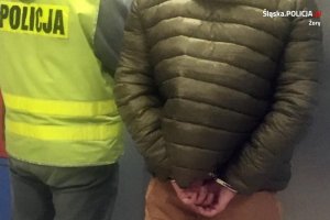 Zatrzymany mężczyzna stoi obok policjanta, ma założone kajdanki