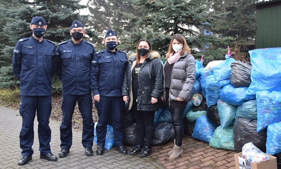 policjanci pracownica cywilna oraz przedstawicielka hospicjum pozują do wspólnego zdjęcia obok worki z uzbieranymi nakrętk