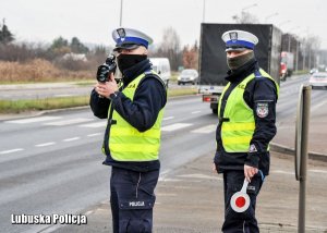 z lewej strony policjant ruchu drogowego trzyma urządzenie do pomiaru prędkości, z prawej strony stoi policjant ruchu drogowego