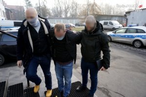 zdjęcie kolorowe - policjanci kryminalni prowadzą zatrzymanego - dwóch policjantów, zatrzymany pomiędzy nimi, w tle samochody w tym radiowozy policji