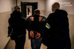 zdjęcie kolorowe - policjanci kryminalni prowadzą zatrzymanego - dwóch policjantów, zatrzymany pomiędzy nimi idą korytarzem