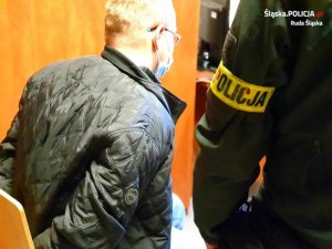 zdjęcie kolorowe - mężczyzna (sprawca) siedzi na krześle obok stoi policjant