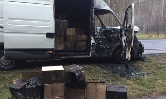Auto dostawcze częściowo spalone. W przestrzeni załadunkowej widoczne przez otwarte drzwi kartony. Kilka kartonów stoi również na poboczu przed autem