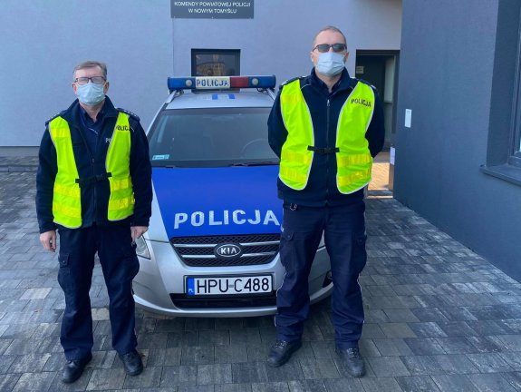 Dwóch umundurowanych policjantów stoi bez czapek, w maseczkach na twarzy. W tle zaparkowany jest oznakowany radiowóz