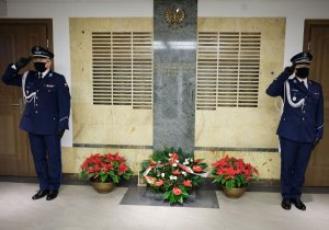 Dwaj policjanci salutują stojąc z dwóch stron Tablicy Pamięci w KGP, symbolicznym miejscem z tabliczkami epitafijnymi z nazwiskami poległych policjantów. Przed Tablicą złożone są kwiaty