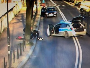 Policyjny radiowóz tarasuje drogę, ma otwarte drzwi. Policjanci udzielają pomocy osobie leżącej na chodniku.