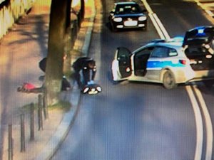 Policyjny radiowóz tarasuje drogę, ma otwarte drzwi. Policjanci udzielają pomocy osobie leżącej na chodniku.