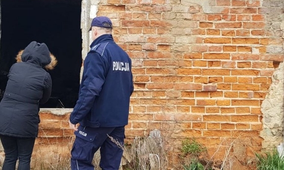 Policjant w mundurze i kobieta w czarnej kurtce zaglądają przez dziurę w ścianie do pustostanu