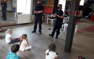 policjanci w maseczkach stoją w sali przedszkolnej zachowując odstęp od siedzących na dywanie dzieci