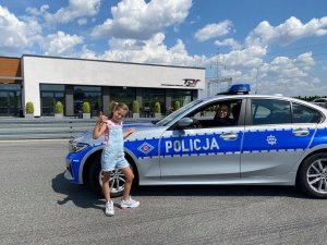 dziewczynka stoi obok radiowozu, w którym siedzi umundurowany policjant