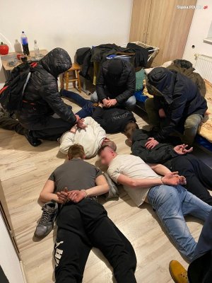 Zatrzymani przez policjantów mężczyźni leżą na podłodze w mieszkaniu, mają na dłoniach kajdanki. Obok nich nieumundurowani policjanci