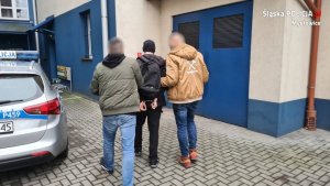 nieumundurowani policjanci prowadzą zatrzymanego przez parking wewnętrzny komendy