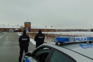 policjanci stoją przy radiowozie, w tle były niemiecki nazistowski obóz koncentracyjny i zagłady Auschwitz – Birkenau