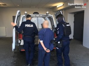 Dwaj umundurowani policjanci wyprowadzają zatrzymanego mężczyznę z radiowozu. Mężczyzna ubrany jest w granatowy dres, niższy niż policjanci i ma kajdanki założone na ręce trzymane z tyłu ciała