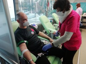 Na zdjęciu pielęgniarka podłącza aparaturę do poboru krwi siedzącemu na fotelu mężczyźnie ubranemu w czarną koszulkę z czerwoną krwinką. Mężczyzna ma zasłonięty nos i usta maseczką ochronną koloru białego