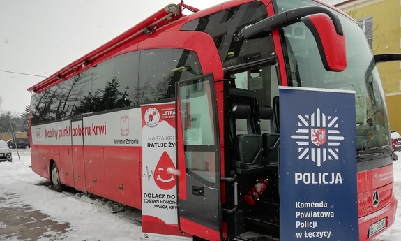 autobus z napisem Mobilny punkt poboru krwi. Po lewej stronie otwartych drzwi usytuowany jest baner koloru biało-czerwonego, na którym widnieje tekst: Nasza krew ratuje życie. Dołącz, zostań honorowym dawcą. HDK PCK Strażak przy OSP w Łęczycy. Po prawej stronie drzwi znajduje się baner koloru granatowego z logo Policji i napisem koloru białego Komenda Powiatowa Policji w Łęczycy