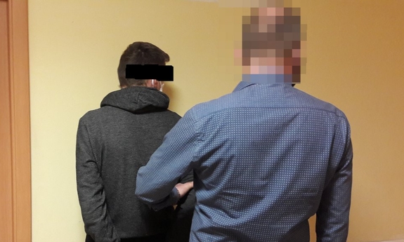 zatrzymany mężczyzna z kajdankami założonymi na przeguby rąk trzymanych z tyłu stoi przy ścianie. Twarzą skierowany do ściany. Po jego prawej stronie za plecami stoi nieumundurowany policjant, który go pilnuje