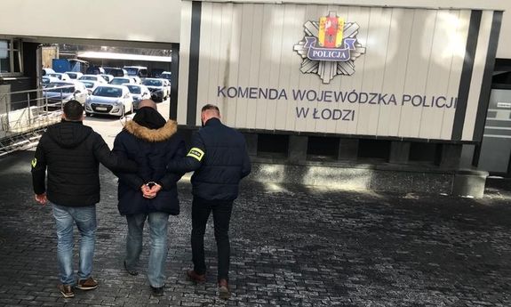 nieumundurowani policjanci konwojują podejrzanego o przestępstwo korupcyjne. Mężczyzna ma założone kajdanki, w tle znajduję się napis Komenda Wojewódzka Policji w Łodzi