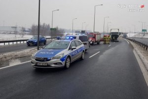 Policyjny radiowóz stoi na drodze przed pojazdem TIR, który brał udział w zdarzeniu drogowym. Na drugim planie pracują policjanci i strażacy