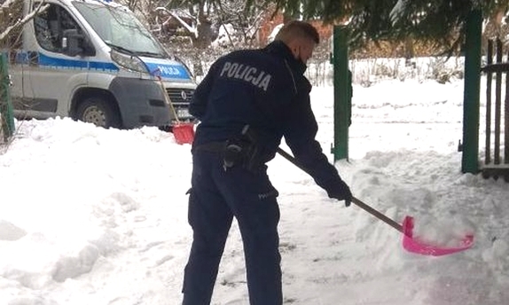 Zdjęcie kolorowe przedstawia posesję w miejscowości Siedliska. Na pierwszym planie widoczny policjant który odśnieża chodnik starszej kobiecie a w tle widoczny radiowóz policyjny