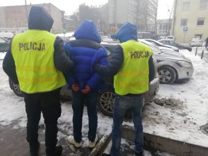 Policjanci ubrani w żółte kamizelki odblaskowe z napisem Policja prowadzą ubranego w niebieską kurtkę zatrzymanego do radiowozu policyjnego. Na trzymane z tyłu ręce mężczyzna ma założone kajdanki