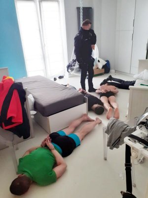 Zdjęcie przedstawia trzech zatrzymanych obywateli Ukrainy podejrzanych o szereg przestępstw, w tym narkotykowych. Trzej mężczyźni leżą na podłoże w szortach oraz t-shirtach. Mają założone kajdanki na ręce z tylu. Nad nimi stoi umundurowany funkcjonariusz Policji.