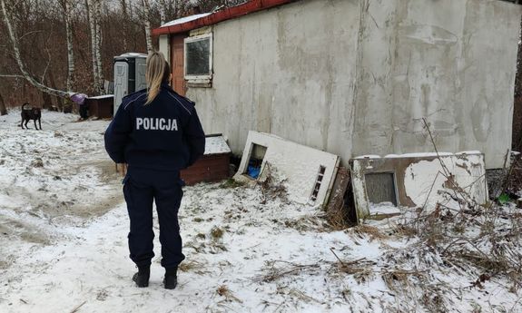 policjantka stojąca tyłem przed nią stara zamieszkała przyczepa cempingową, toi toi oraz biegający pies