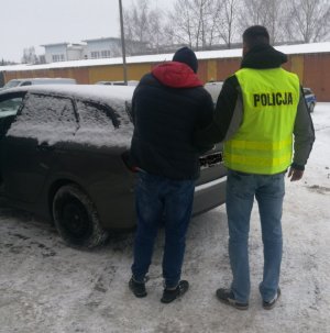 z tyłu budynku komendy stoi nieoznakowany radiowóz przed którym stoi policjant ubrany w kamizelkę odblaskową  z napisem policja, i trzyma zatrzymanego, który ma założone kajdanki na ręce trzymane z przodu oraz na stopy, w tyle budynki gospodarcze