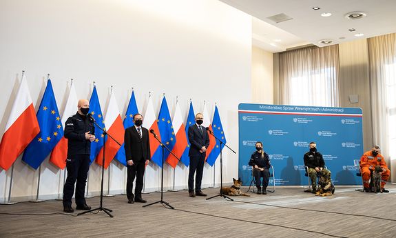 Przed mikrofonami podczas konferencji prasowej stoją od lewej: Komendant Główny Policji gen.insp. Jarosław Szymczyk w mundurze ćwiczebnym, Minister SWiA Mariusz Kamiński w garniturze i Wiceminister SWiA Błażej Poboży. W tle widoczne sa flagi UE i Polski. Z prawej strony na krzesłach siedzą opiekunowie psów służbowych Policji, Straży Granicznej i Państwowej Straży Pożarnej wraz ze swymi podopiecznymi. Za ich plecami widoczna jest ścianka z napisami: Ministerstwo Spraw Wewnętrznych i Administracji.