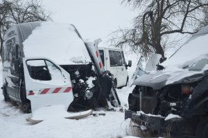 Na zdjęciu widoczne są trzy pojazdy typu bus, dwa koloru czarnego są zasypane śniegiem i posiadają uszkodzone i zdekompletowane przody. Na jednym z czarnych busów oparte są drzwi koloru białego z biało czerwoną naklejką. W oddali widać tylko część białego busa, który stoi za czarnymi busami