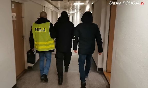 Dwaj policjanci ubrani po cywilnemu, jeden w żółtej kamizelce z napisem Policja na plecach, prowadzą zatrzymanego mężczyznę