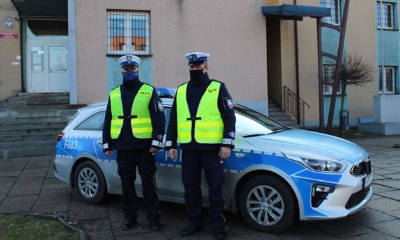 Zdjęcie przedstawiające parking przed wieluńską komendą Policji i zaparkowany przed budynkiem radiowóz policyjny. Przed radiowozem stoją dwaj umundurowani policjanci drogówki