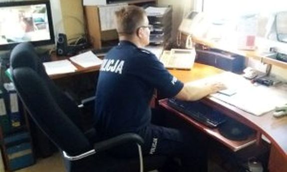 policjant służby dyżurnej w koszulce z napisem Policja na plecach przy swoim stanowisku pracy. Przy nim stoi komputer i widoczne są telefony