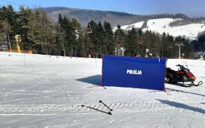 ośnieżony stok, na nim rozłożony granatowy parawan kryminalistyczny z napisem Policja,  z boku widać skuter śnieżny, rozrzucone kijki narciarskie, w tle wyciąg