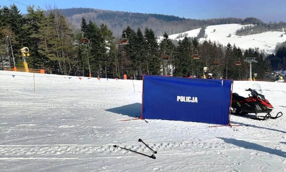 ośnieżony stok, na nim rozłożony granatowy parawan kryminalistyczny z napisem Policja,  z boku widać skuter śnieżny, rozrzucone kijki narciarskie, w tle wyciąg