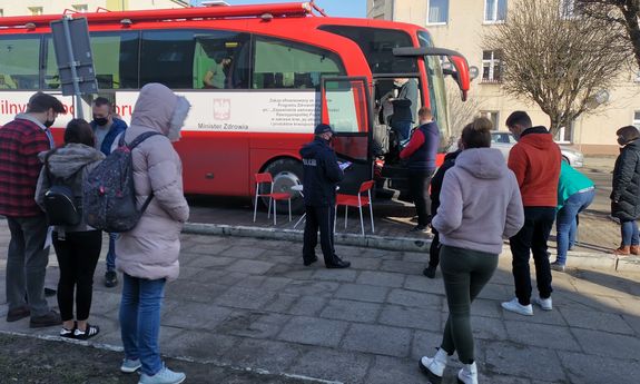 mieszkańcy oraz policjant stoją w kolejce przez autobusem służącym do oddania krwi