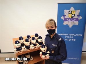 Policjantka trzyma Misia - maskotkę, z boku ustawione są inne miśki, a z tyłu widoczny baner z logo Komendy Powiatowej Policji w Sulęcinie