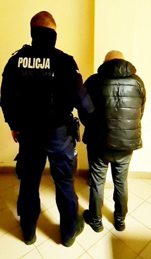 Po lewej stronie tyłem stoi policjant w umundurowaniu służbowym, który trzyma za przedramię stojącego po prawej stronie zatrzymanego, który również jest odwrócony tyłem.&quot;&gt;