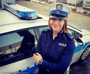 policjantka w granatowym mundurze, białej czapce na głowie i białym pasie na biodrach stoi przy oznakowanym radiowozie&quot;&gt;