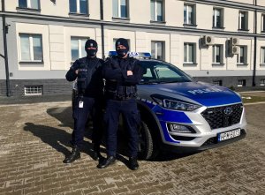 Dwóch umundurowanych policjantów stoi przy oznakowanym radiowozie. W tle budynek Komendy Powiatowej Policji w Kolnie.&quot;&gt;