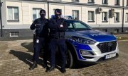 Dwóch umundurowanych policjantów stoi przy oznakowanym radiowozie. W tle budynek Komendy Powiatowej Policji w Kolnie.&quot;&gt;