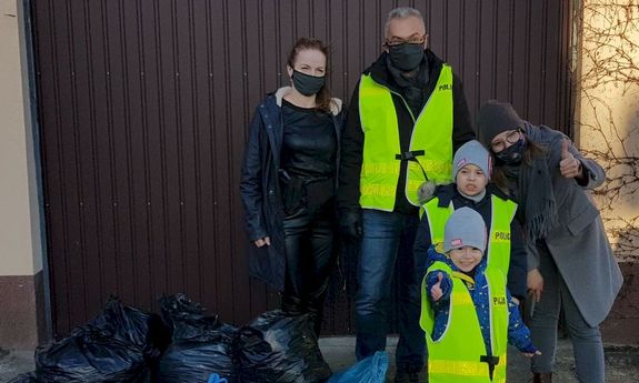 Dwie kobiety, mężczyzna ubrany w żółtą kamizelkę z napisem Policja, dwójka dzieci w żółtych kamizelkach, obok widoczne foliowe worki wypełnione nakrętkami